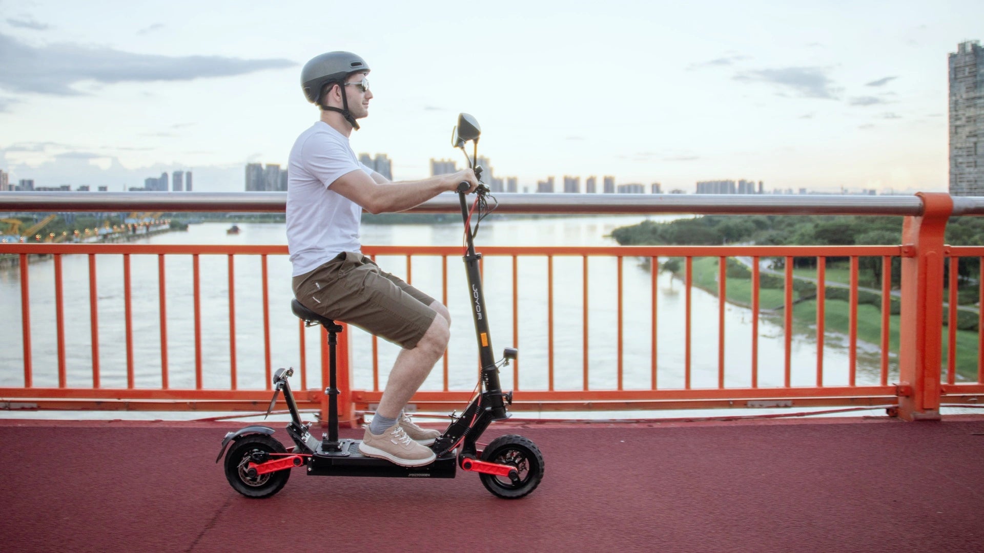 Video laden: City commuting footage of Joyor S8E EU legal seated electric scooter, showing comfortable seat, foldable design and smooth riding along river urban bridge